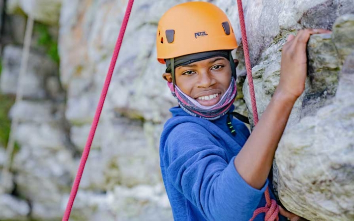 a young person wearing a helmet and secured by ropes pauses rock climbing to smile at the camera